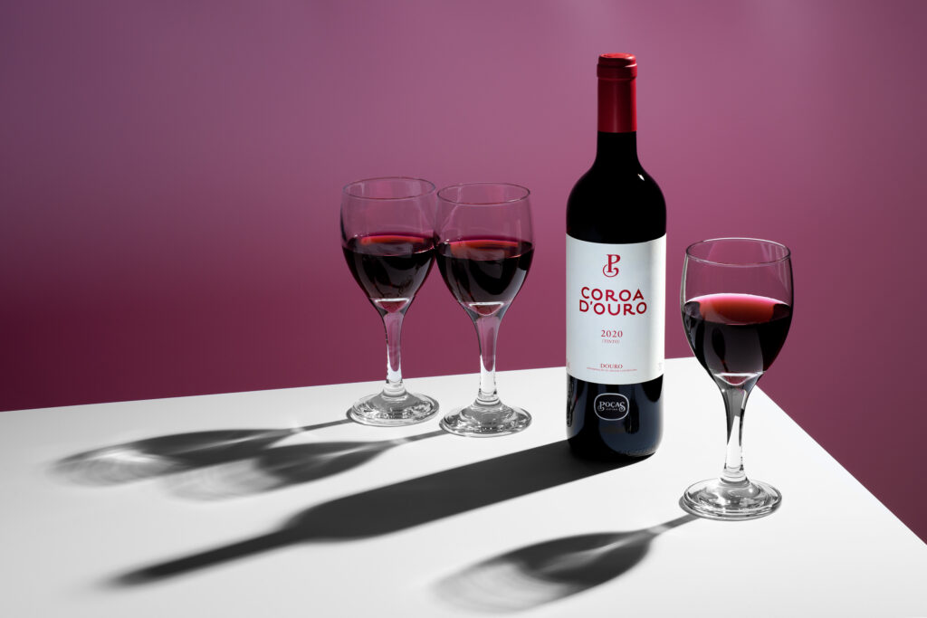 Bouteille de vin Coroa d’Ouro et trois verres remplis projetant de fortes ombres sur un fond rose