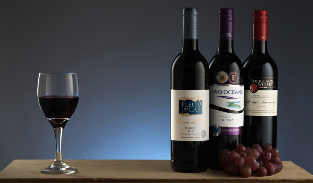 Three bottles of red wine with a filled wine glass and grapes on a table